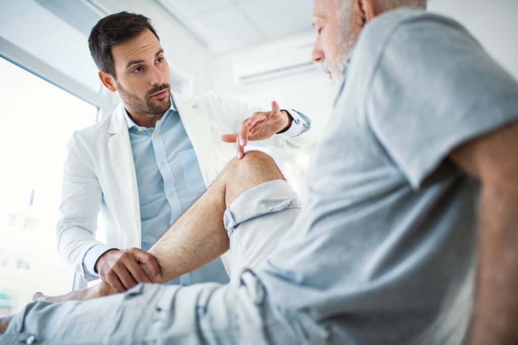 Closeup side view of early 30's doctor examining a knee of a senior gentleman during an appointment. The doctor is gently touching the tendons around the knee and showing the patient which part of his knee has suffered.