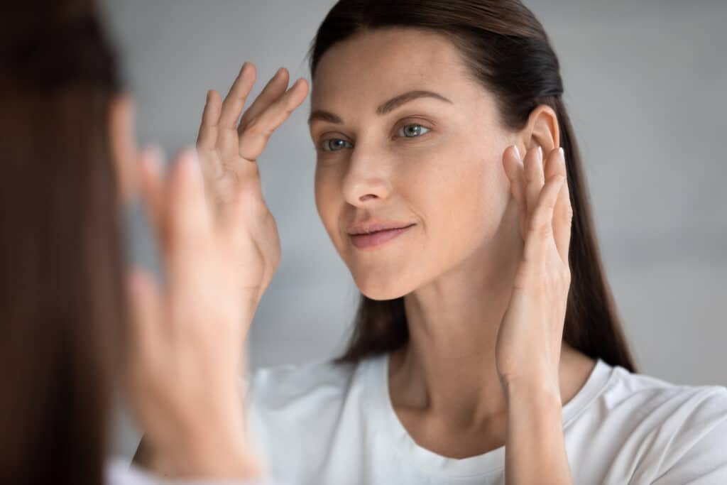 woman examining her facial skin in a mirror while considering moxi, halo, or co2 laser resurfacing treatments.